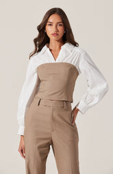 A woman wearing a white shirt with long, puffed sleeves and buttoned cuffs, layered with a beige strapless bustier top over the shirt. She is also dressed in matching beige high-waisted trousers with belt loops and a front zipper.